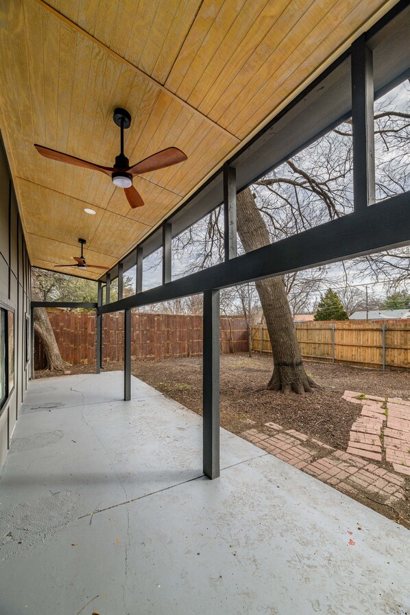 An outdoor covered patio with a ceiling fan, large windows, and a view of a backyard with a tree and a wooden fence. - Dallas Luxe Retreat- Lavish 4-Bedroom Escape (Dallas)