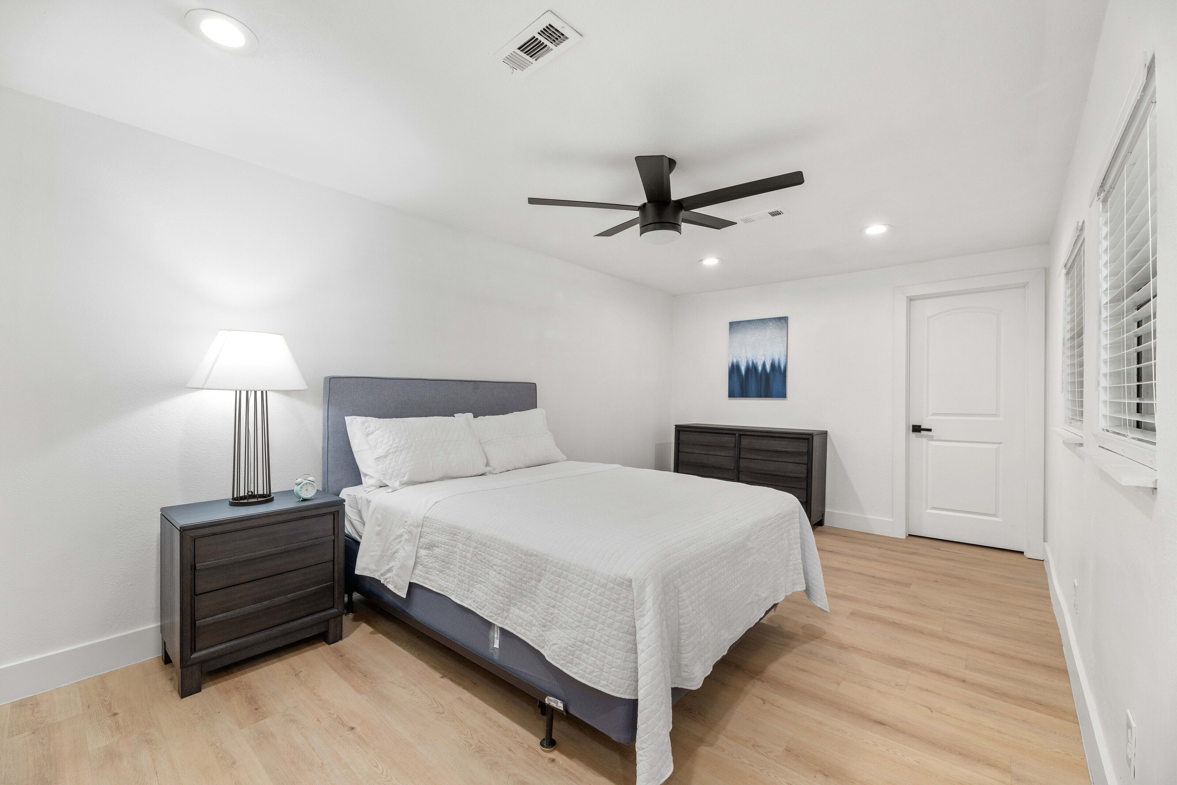 A bedroom with a bed, nightstands, a ceiling fan, a wall-mounted lamp, a door, and a window with blinds.
