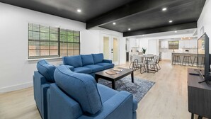 A modern living room with a blue sectional sofa, a coffee table, and a TV on a stand. There is a dining area with a table and chairs, and a kitchen in the background. - Forest Hill Family Hideaway- 4-Bed Retreat (Dallas)