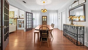 Ein Esszimmer mit einem Holztisch, Stühlen und einer Chandelüster. An den Wänden befinden sich ein Bücherregal und ein Spiegel.