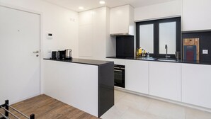 A modern kitchen with white cabinetry, a black countertop, and a built-in oven. There is a window with a black frame and a wooden cutting board on the counter. - GuestReady - comfort near Jardim do Morro (Vila Nova de Gaia)