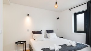 A neatly made bed with white bedding and two pillows, a small black side table, and a single hanging light bulb. - GuestReady - comfort near Jardim do Morro (Vila Nova de Gaia)