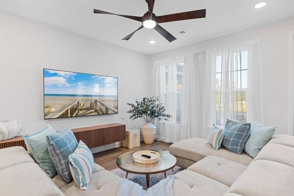 A living room with a ceiling fan, a large beach-themed painting, a sofa set, a round coffee table, and a potted plant.