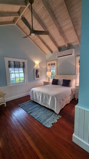 A bedroom with a wooden ceiling, a large bed, a window with a view, and an air conditioning unit.