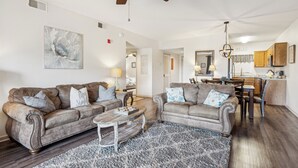Un salon avec deux canapés, une table basse et un tapis. On y trouve également un coin repas avec une table et des chaises. Un ventilateur de plafond est visible, et les couleurs de la pièce sont neutres.
