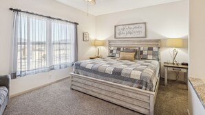 Une chambre à coucher avec un lit, un ventilateur de plafond, une table de chevet avec une lampe, une fenêtre avec des rideaux légers et une citation inspirante sur le mur.
