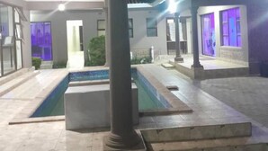 A modern outdoor pool area with a covered seating area and a well-lit building in the background.