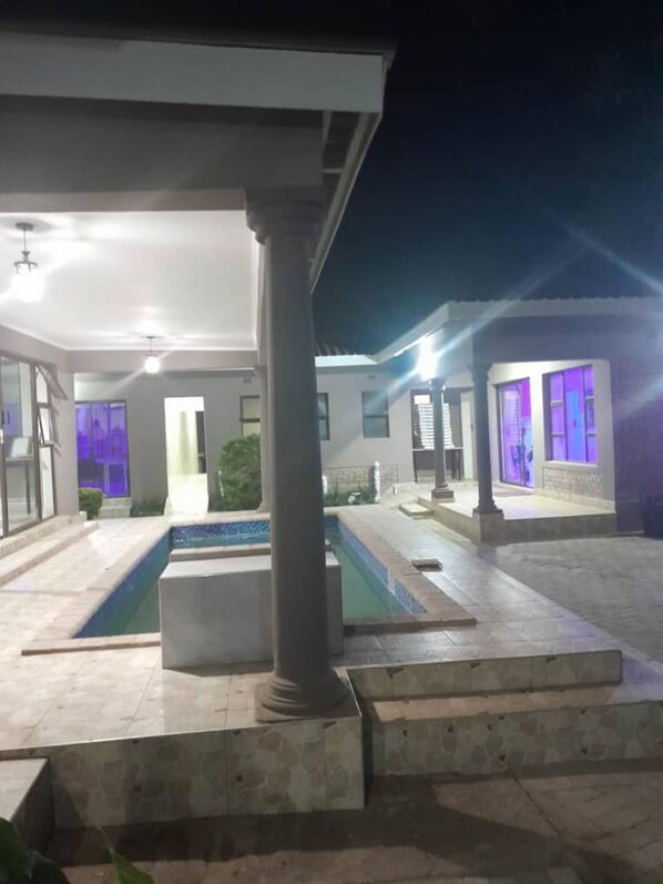 A modern outdoor pool area with a covered seating area and a well-lit building in the background. - Modernly Furnished Standard Room With Queen bed - 2220 (Kadoma)
