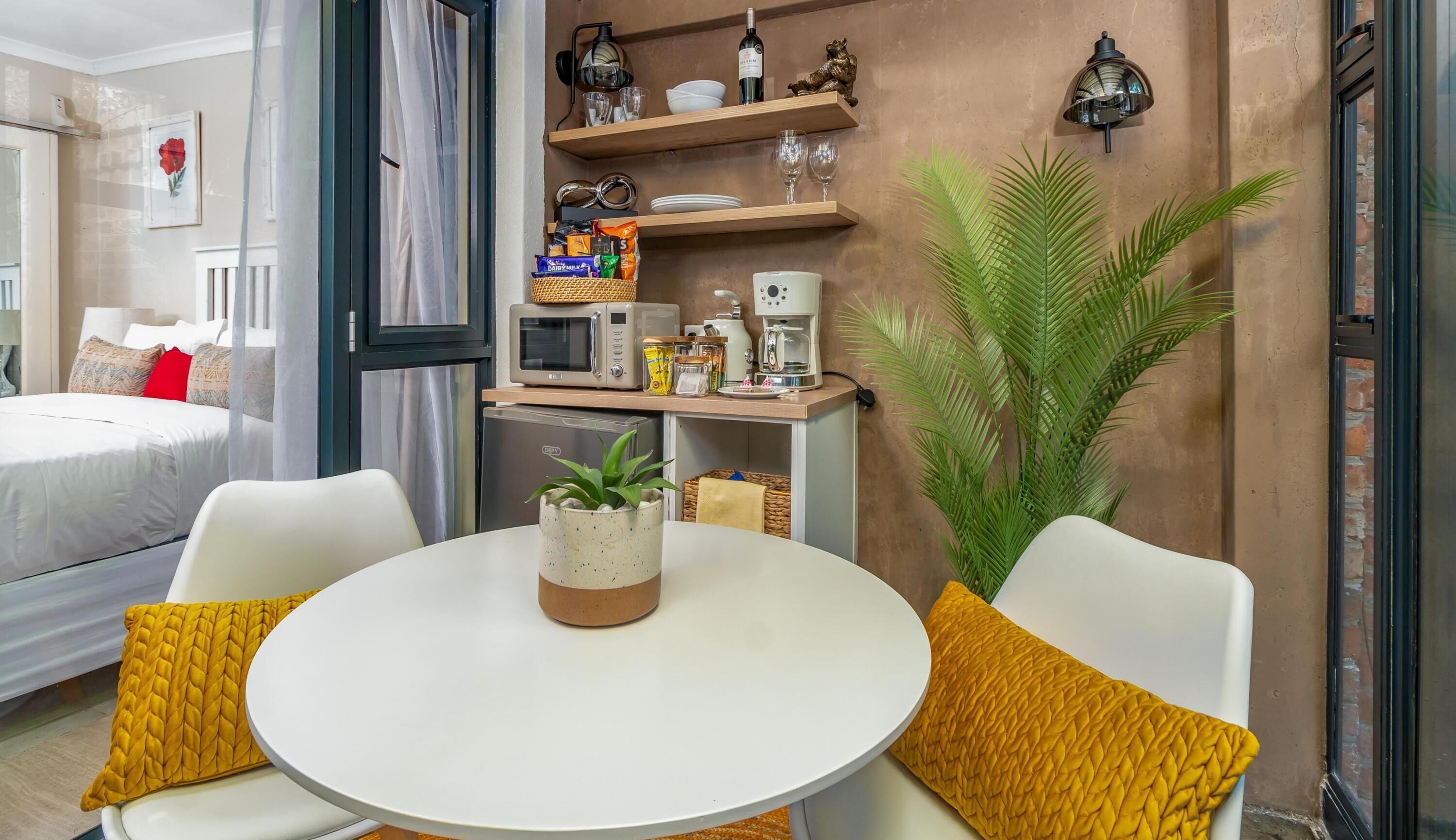A hotel room with a bed, a round table with two chairs, a coffee machine, a microwave, and a small potted plant.