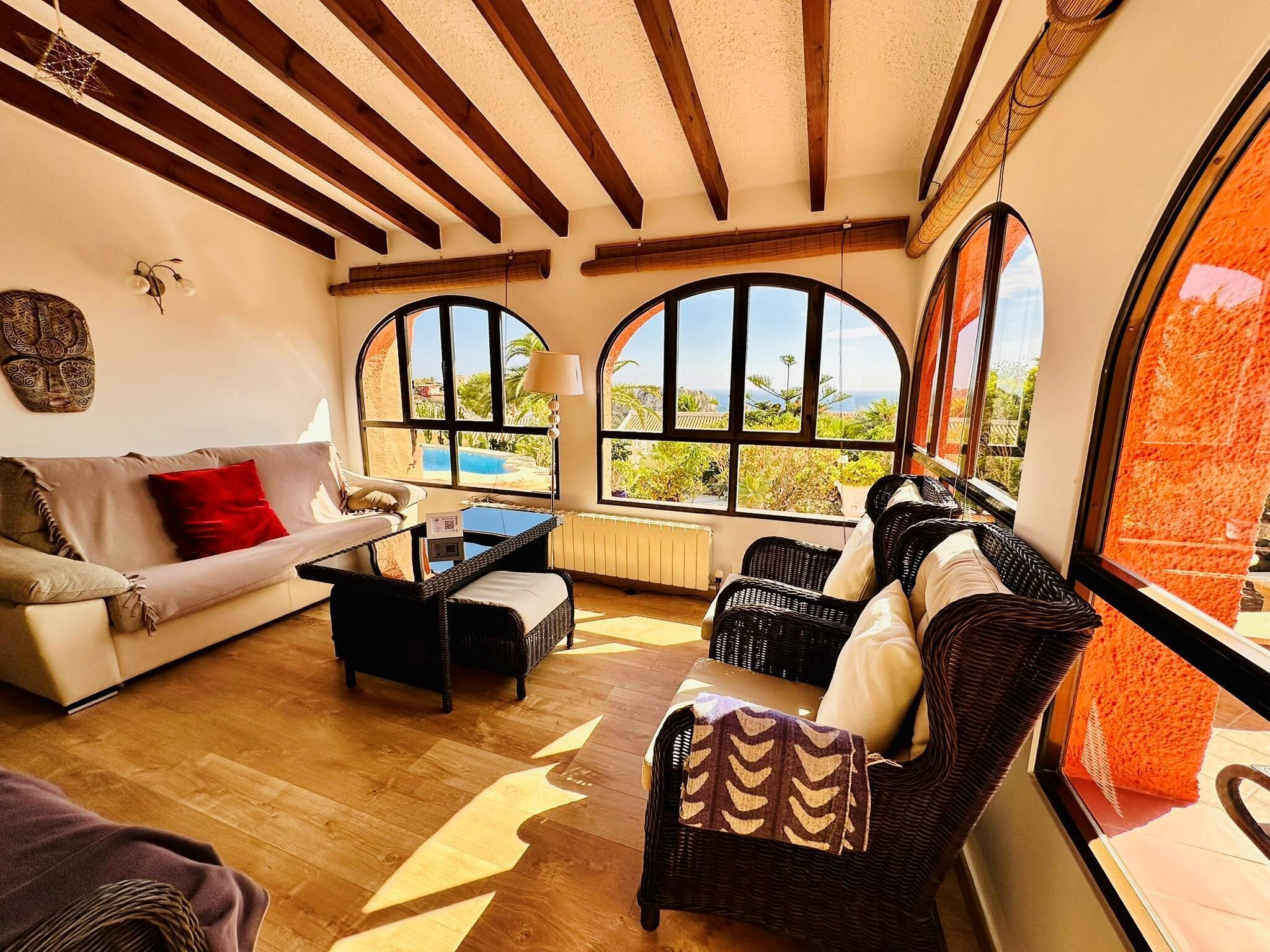 A cozy living room with a sofa, a coffee table, and wicker chairs. Large arched windows offer a view of a swimming pool and greenery.