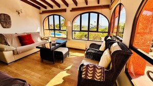 A cozy living room with a sofa, a coffee table, and wicker chairs. Large arched windows offer a view of a swimming pool and greenery.