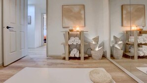 A hallway with a wooden floor, a white door, and a mirror reflecting the room. There are shelves with folded towels and decorative items. - Wilderness Ranch Retreat (Boise)