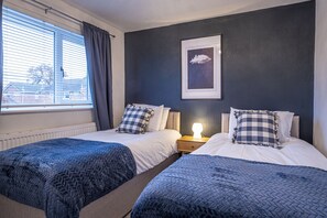 A bedroom with two beds, a window with blinds, a framed picture on the wall, and a bedside table with a lamp. - Stylish 2 bedroom house in quiet suburb of Chester Sleeps 6 by Restfully (Blacon)