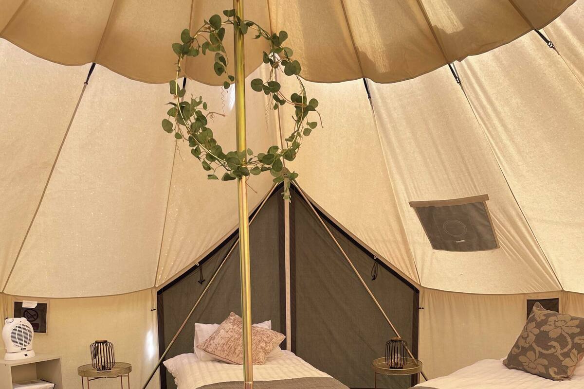Ein Zelt mit drei Betten, jedes mit einer grauen Decke und Kissen, einem kleinen Tisch mit Stuhl und einer Zimmerpflanze.