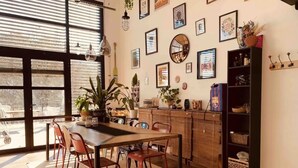Un comedor con una mesa y sillas de madera, un ventanal con persianas, y una pared decorada con varias imágenes enmarcadas y un espejo.