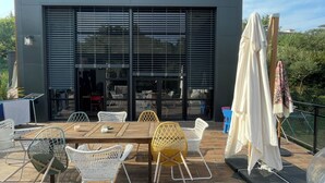 Un patio exterior con un edificio moderno al fondo, que incluye una mesa y sillas, y una zona de estar cubierta.