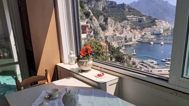A balcony with a table set for tea, overlooking a coastal town with buildings and a marina. - Apartament in Amalfi ID 4178 (Amalfi)