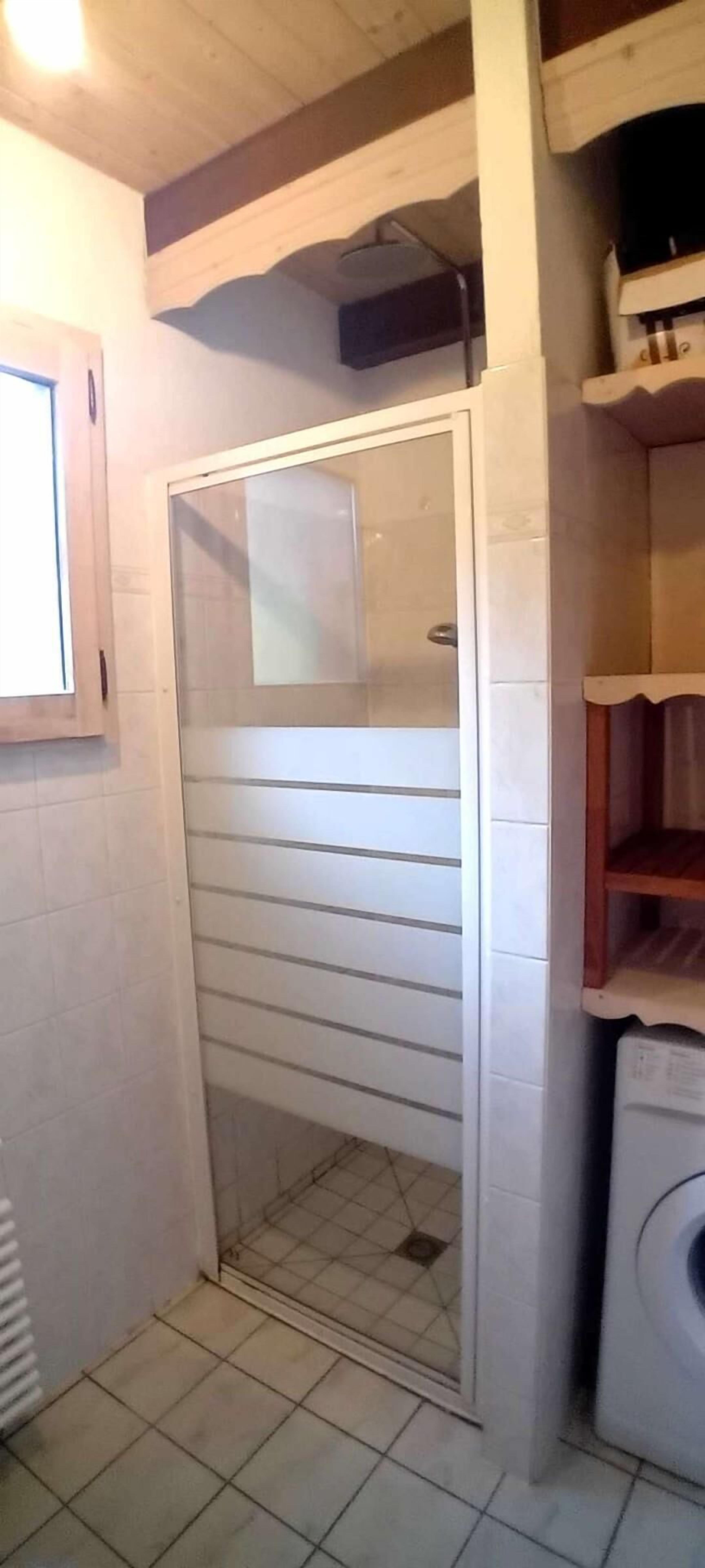 Een kleine, afgesloten badkamer met een douche, een raam en een wasmachine.