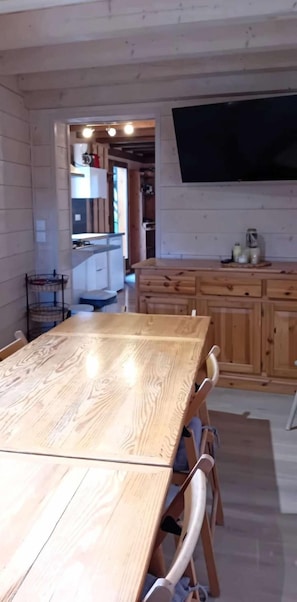 A wooden dining table with chairs, a kitchen area with cabinets, and a flat-screen TV mounted on the wall.
