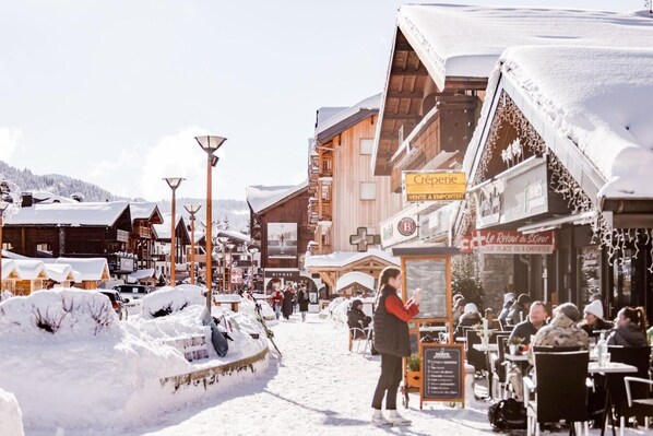 A snowy village scene with wooden buildings, outdoor seating, and a sign for 'Creperie'.