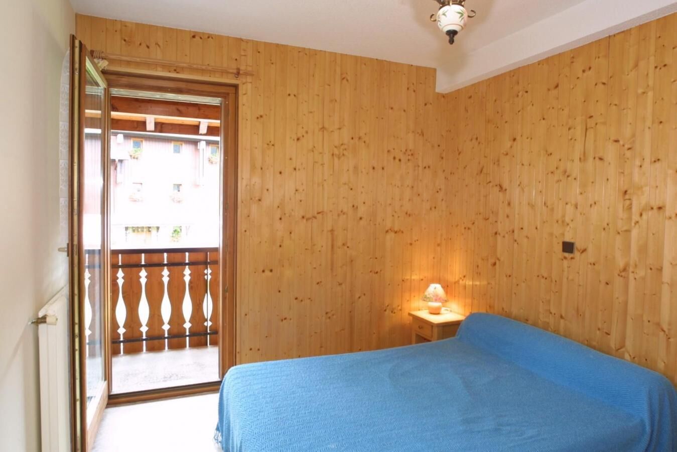 A room with wooden panel walls, a bed with a blue cover, a nightstand with a lamp, and a balcony door.
