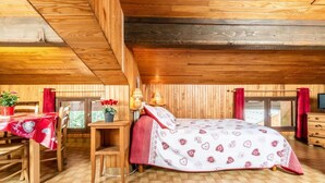 Un dormitorio acogedor en el ático con techo de madera, una cama con ropa de cama estampada, un escritorio con silla y una ventana con cortinas.