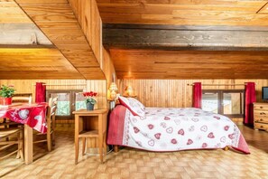 Une chambre mansardée chaleureuse avec un plafond en bois, un lit recouvert d’une literie à motifs, un bureau avec une chaise et une fenêtre avec des rideaux.