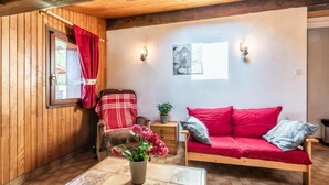 Una habitación acogedora con paredes de madera, un sofá rojo, una silla de madera, una mesita con un jarrón de flores y una ventana con cortinas rojas.