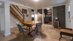 Un coin repas avec une table et des chaises en bois, un escalier et une cuisine visible à travers une porte ouverte.