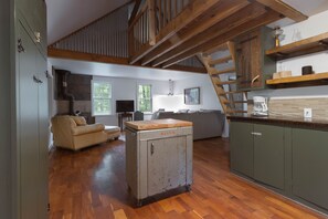 A spacious living area with a wooden floor, a sofa, a kitchenette with a coffee maker, and a staircase. - Quaint cottage in the woods with swim spa (Frazeysburg)