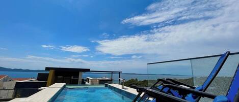 A rooftop pool with clear blue water, surrounded by a glass balustrade, and overlooking a scenic landscape with mountains in the distance.