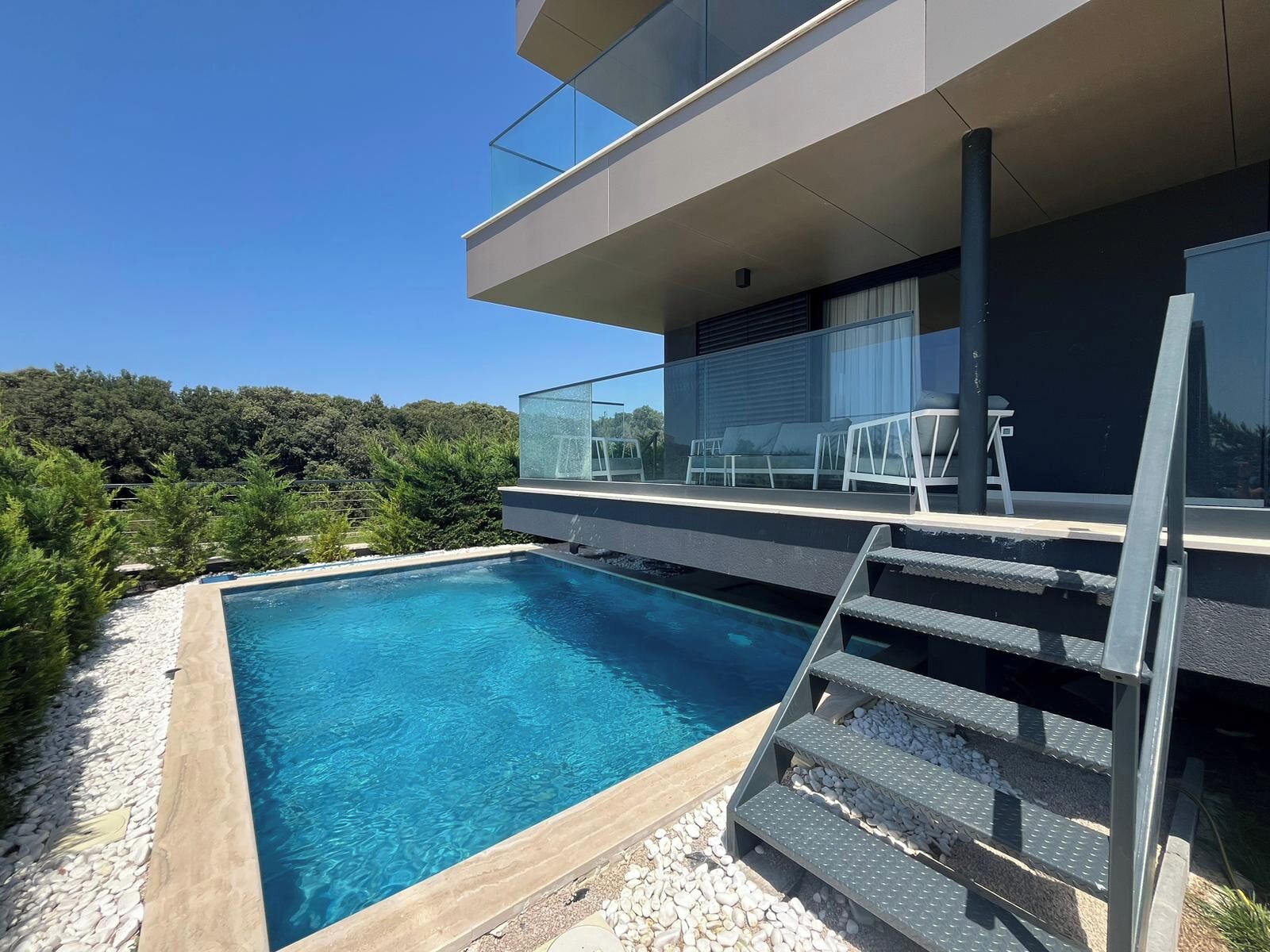 Ein moderner Poolbereich mit einem klaren, blauen Pool, Stufen, die zu einem Balkon führen, und Blick auf einen bewaldeten Hang.