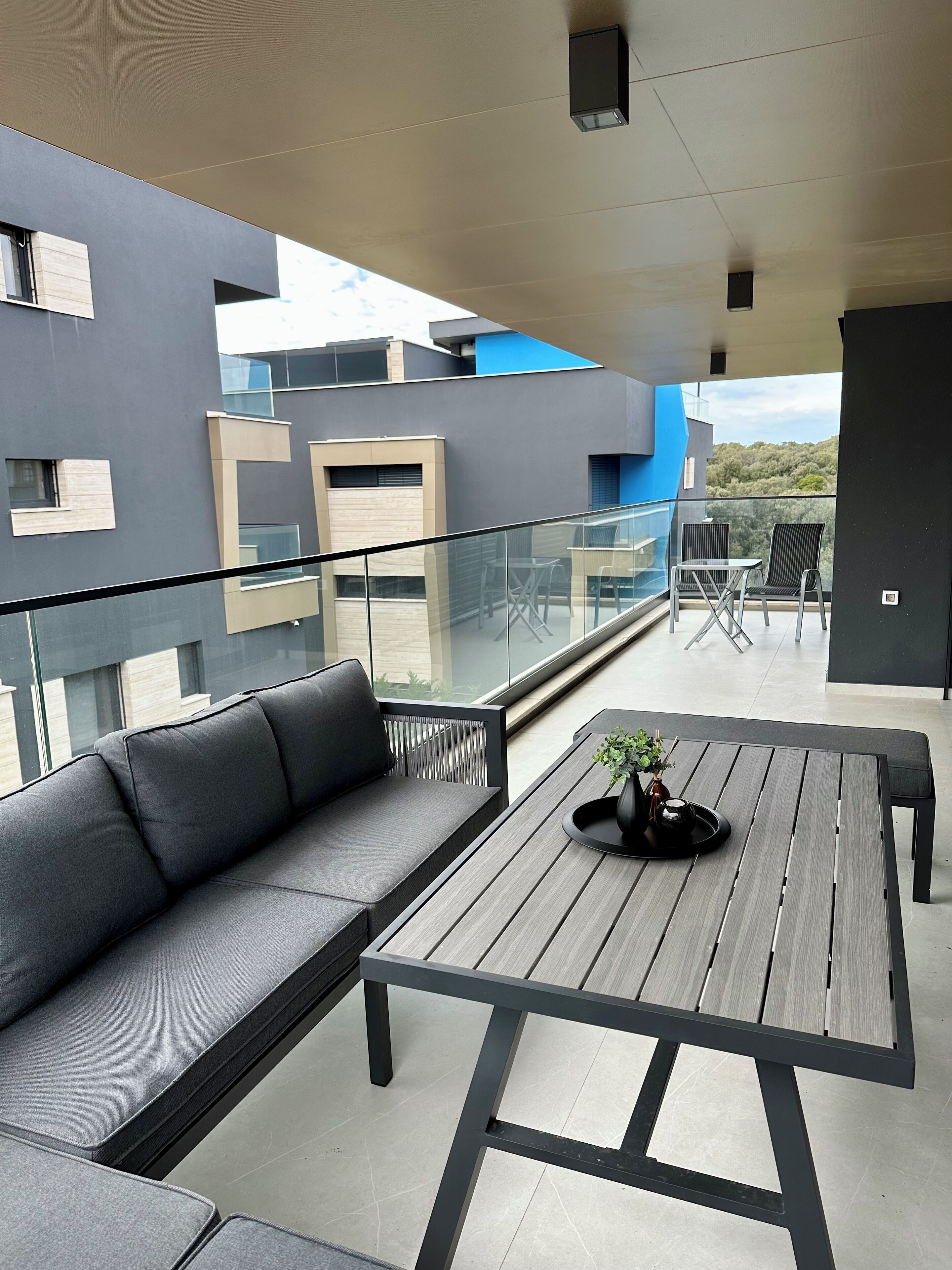 A modern balcony with a grey sofa and a wooden table, featuring a potted plant.