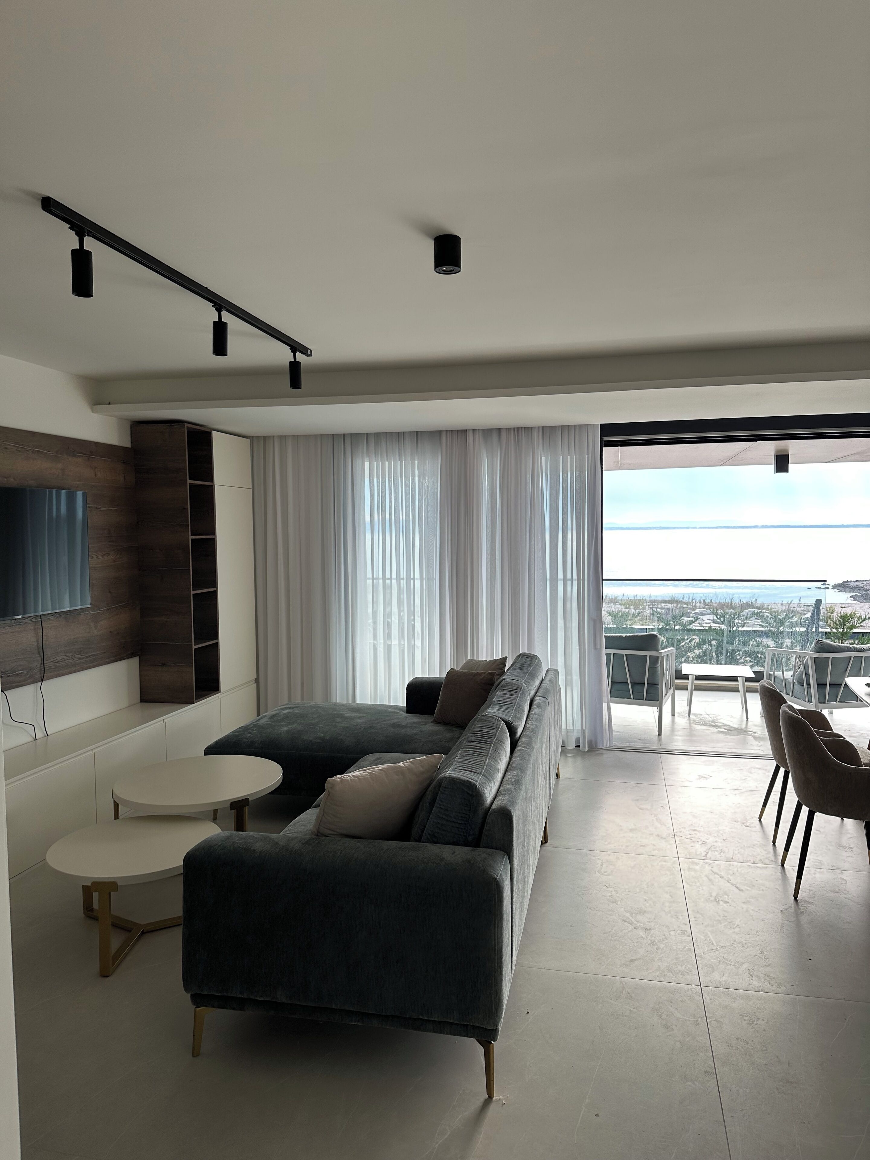A modern living room with a sectional sofa, a flat-screen TV mounted on a wooden wall, a dining area with a table and chairs, and a large window offering a city view.
