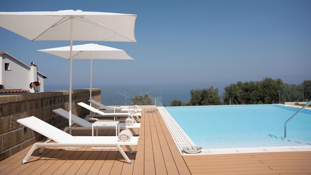 A poolside area with white sun loungers and umbrellas, a clear view of the sea, and a wooden deck. - Tenuta Sirignano Villa Nord e Sud (Massa Lubrense)