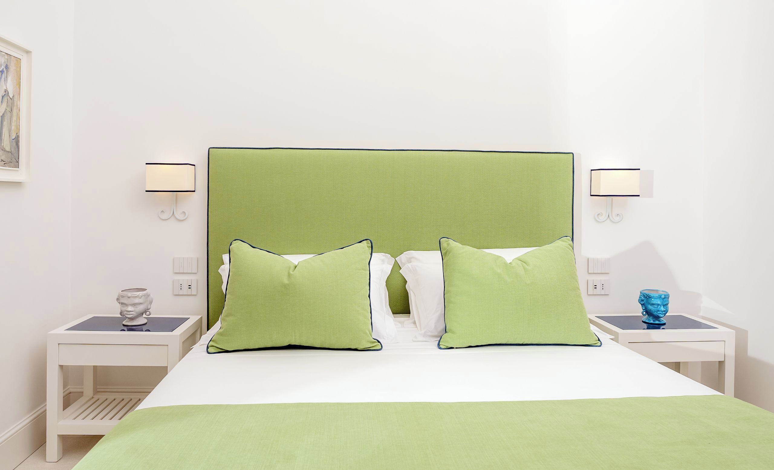 Habitación de hotel ordenada con una cama grande, almohadas verdes, ropa de cama blanca y dos mesitas de noche con lámparas y objetos decorativos.