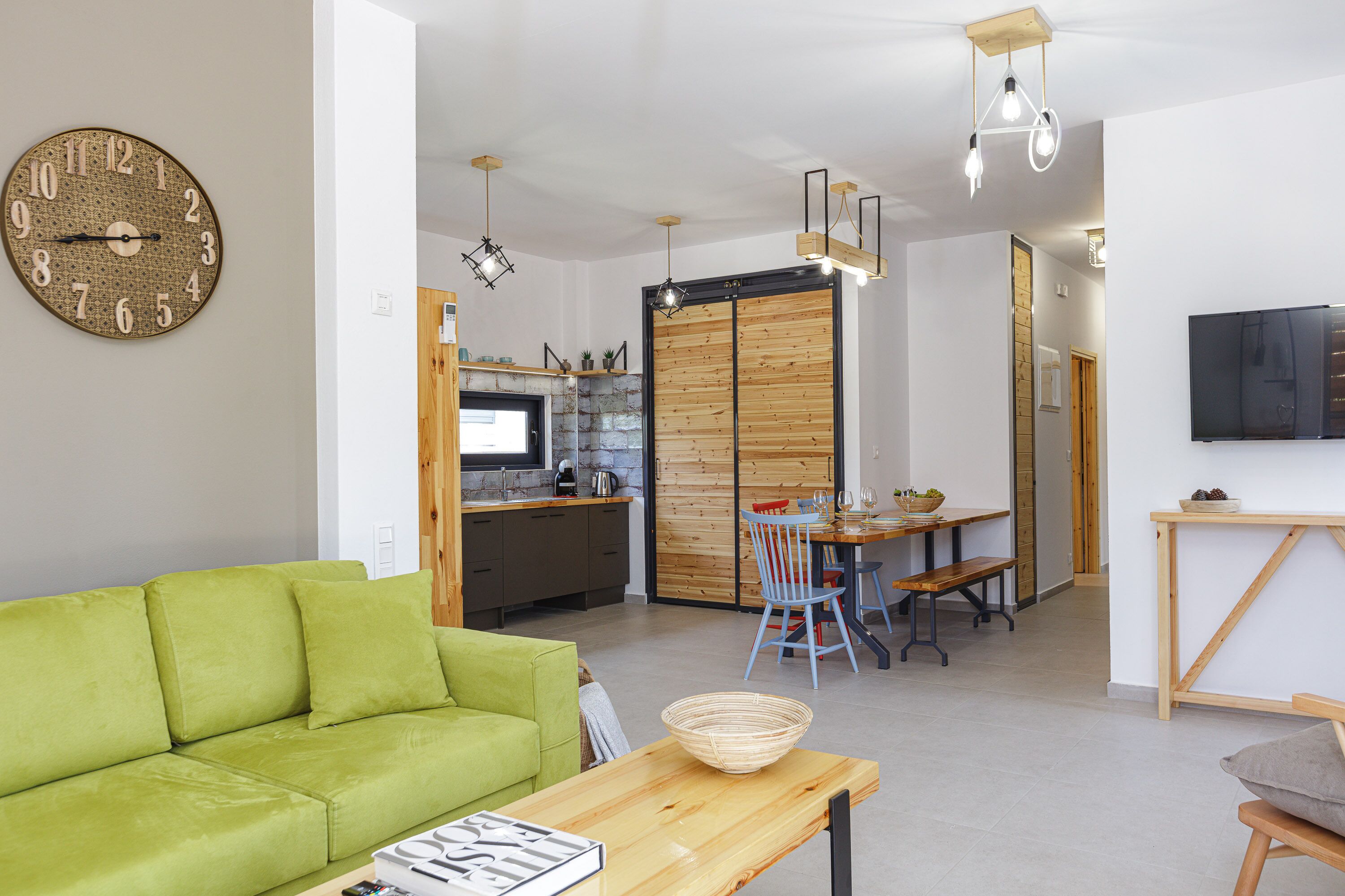 A modern living space with a green sofa, wooden dining table, and a kitchen area with a TV and wooden cabinets.