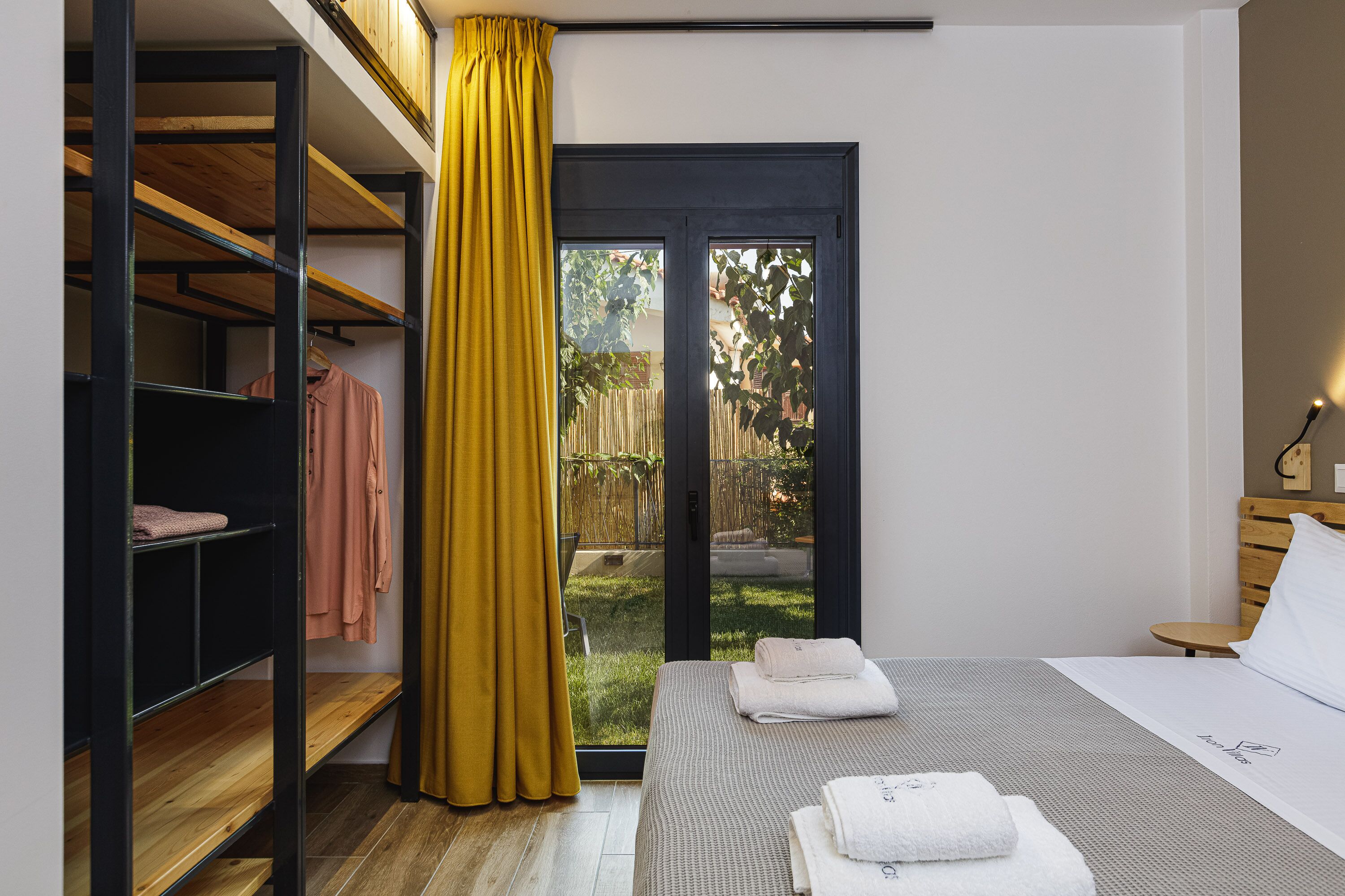 Un dormitorio con una cama, una estantería de madera, una cortina amarilla y vistas a un jardín a través de un ventanal.