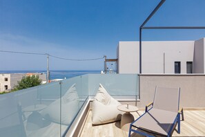 A balcony with a glass railing, a lounge chair, a small table, and a cushion. - Iron Villas Crete (Rethymno)