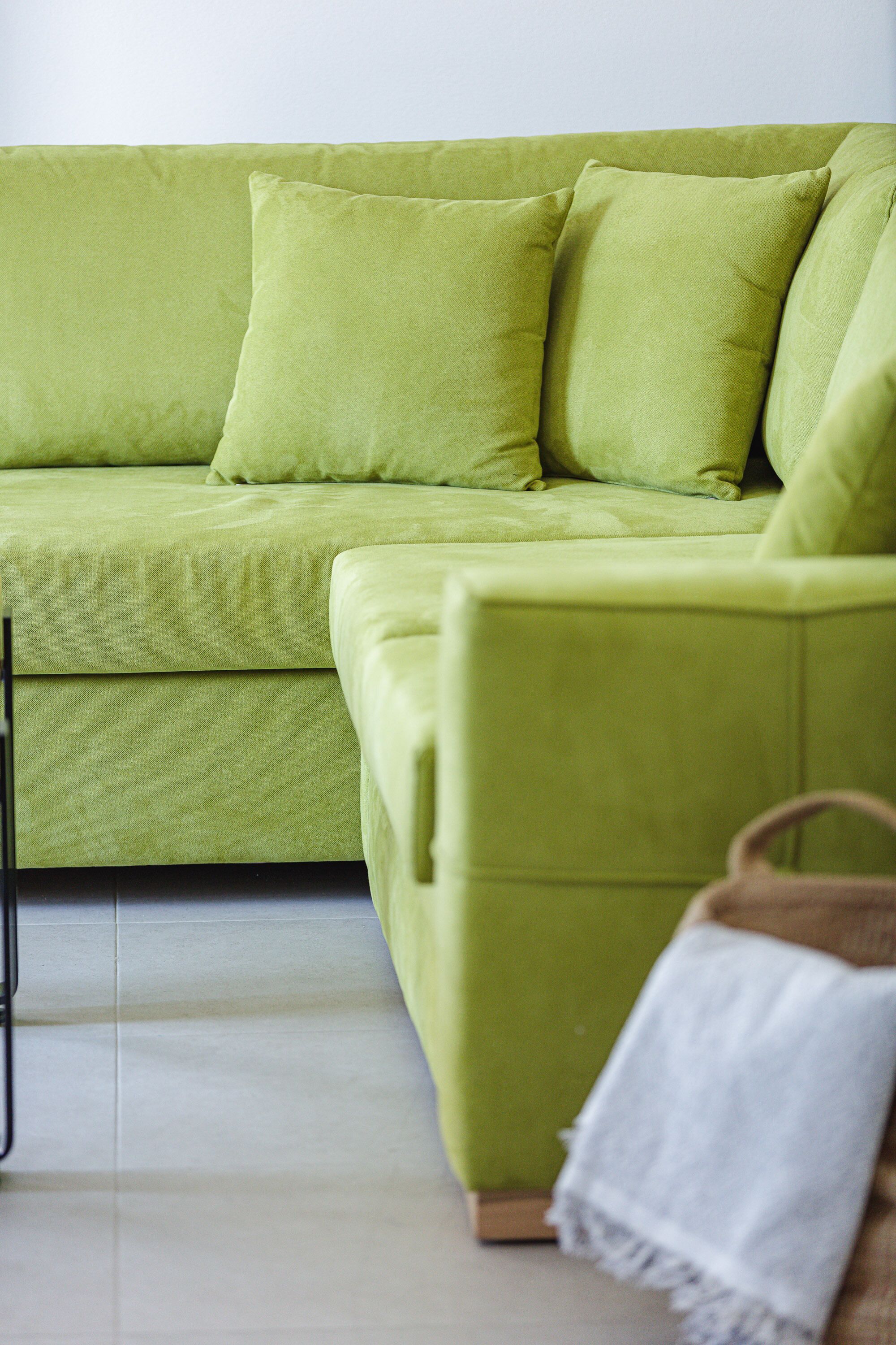 A green sofa with multiple cushions, a white throw blanket, and a wooden chair in a room with tiled flooring.
