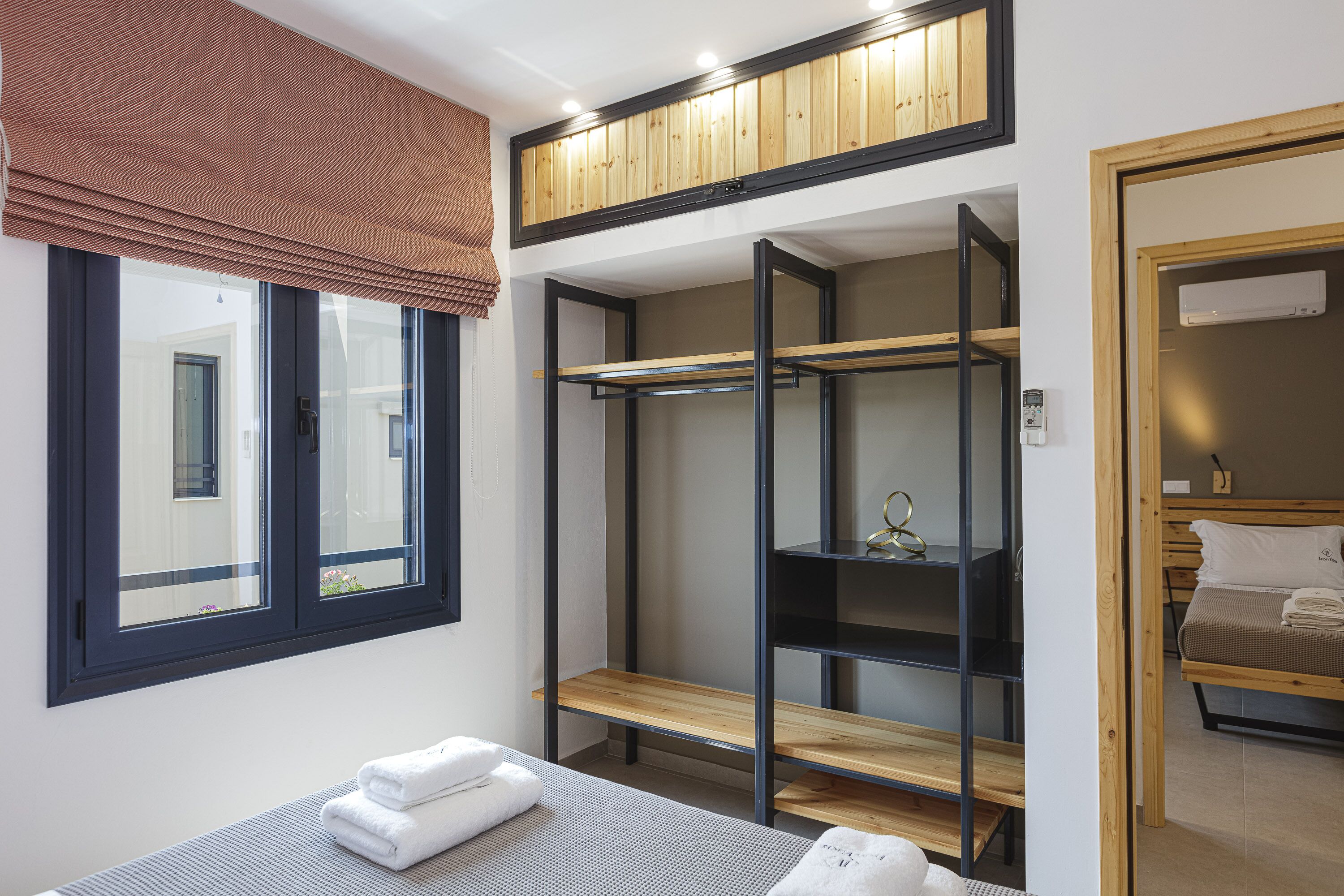 A bedroom with a bunk bed, a wooden shelf unit, a window with blinds, and a bedside table with towels.