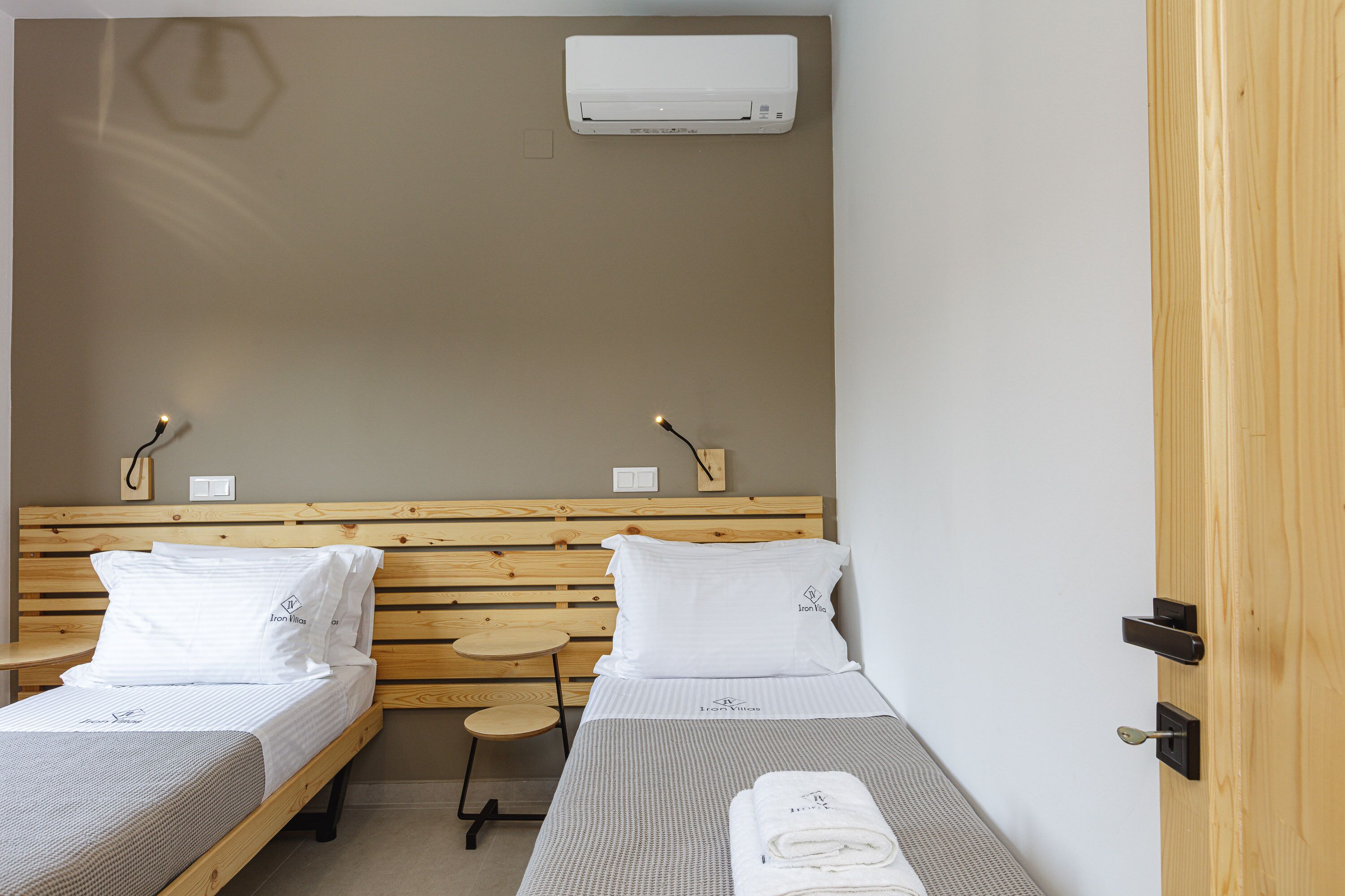 Een kamer met twee enkele bedden, elk met een houten hoofdbord en wit beddengoed. Daartussen staat een klein houten krukje, en boven de bedden is een airconditioning aangebracht.