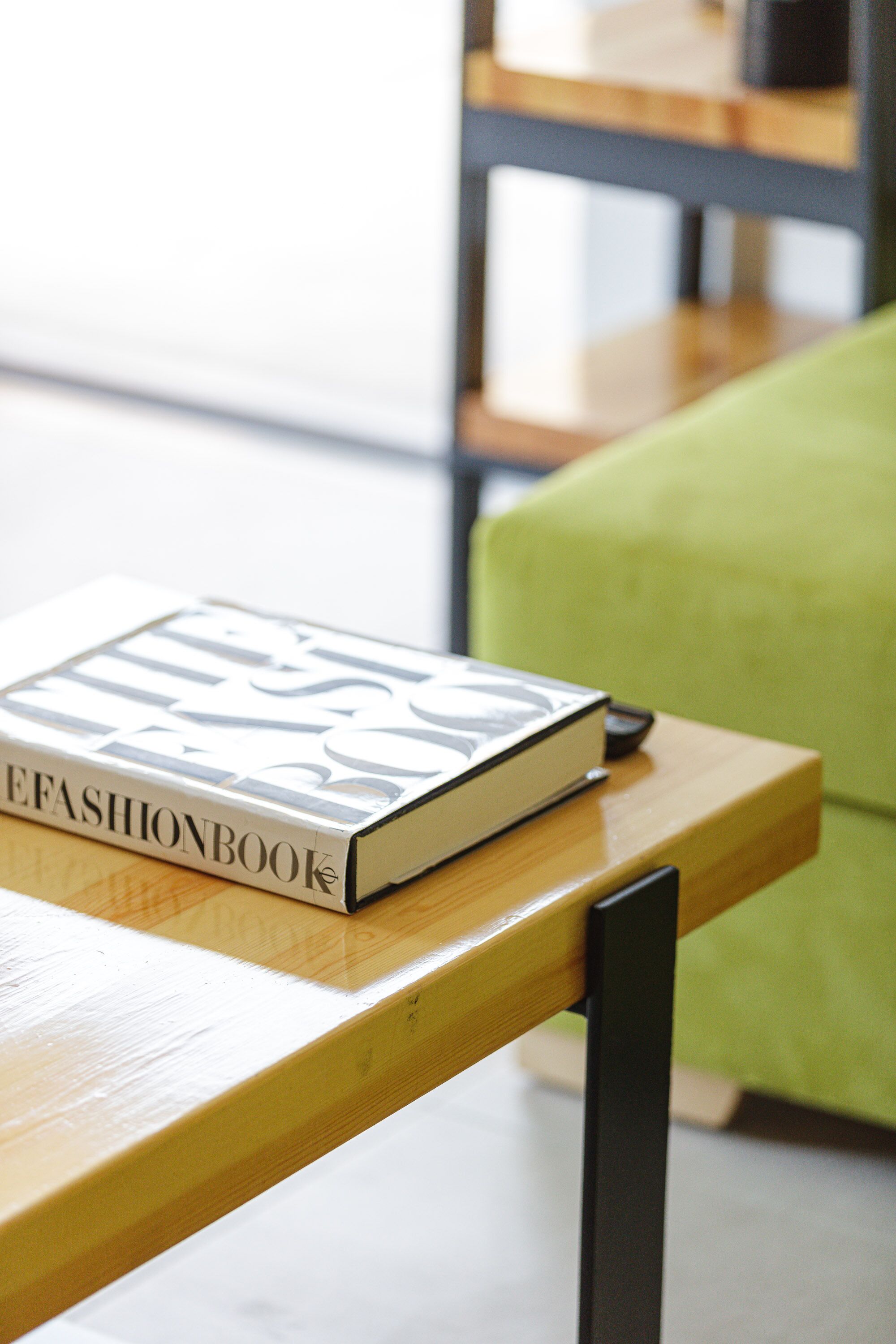 A book titled "FASHIONBOOK" on a wooden table with a green chair in the background.
