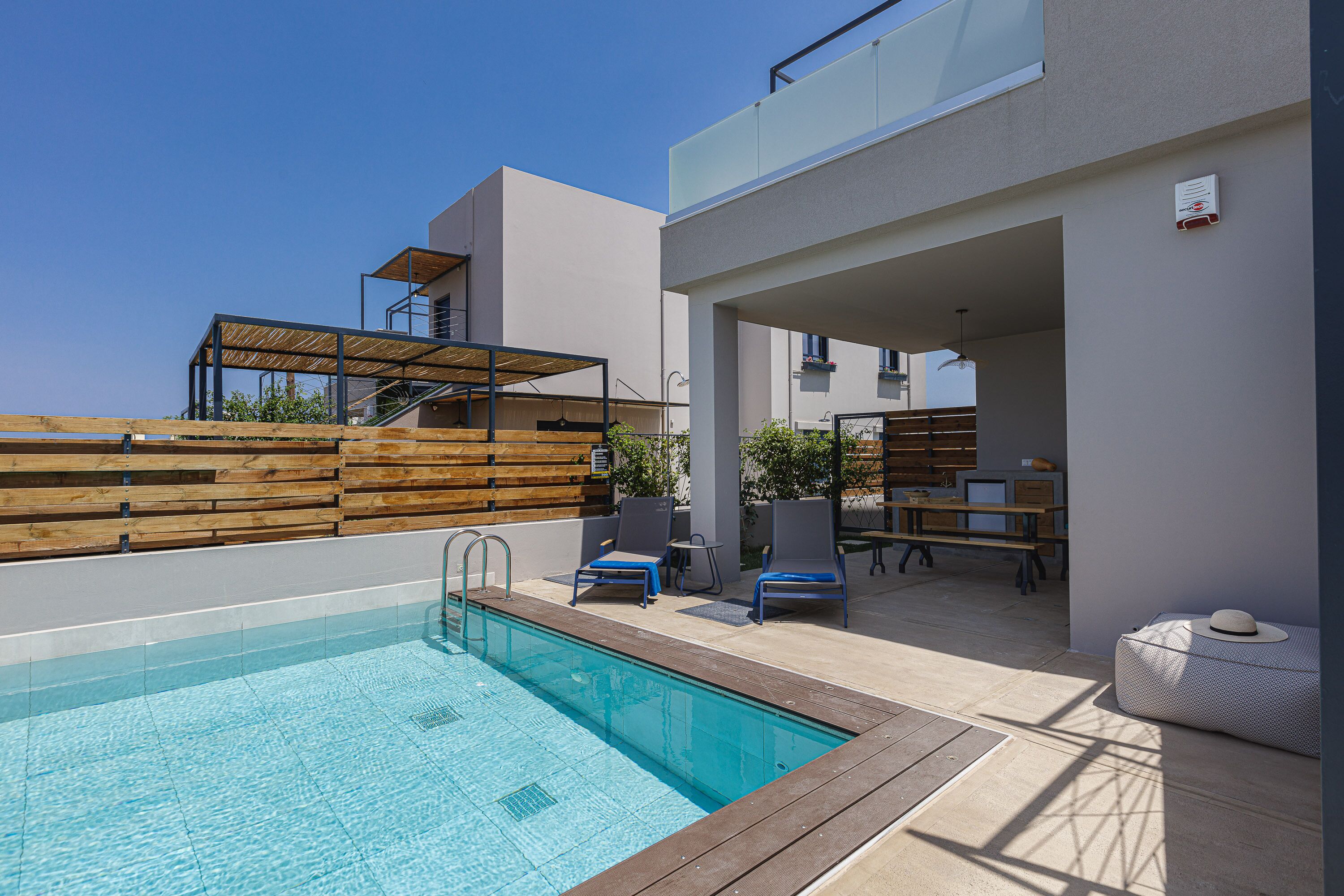 A modern outdoor pool area with a wooden deck, lounge chairs, and a covered seating area.
