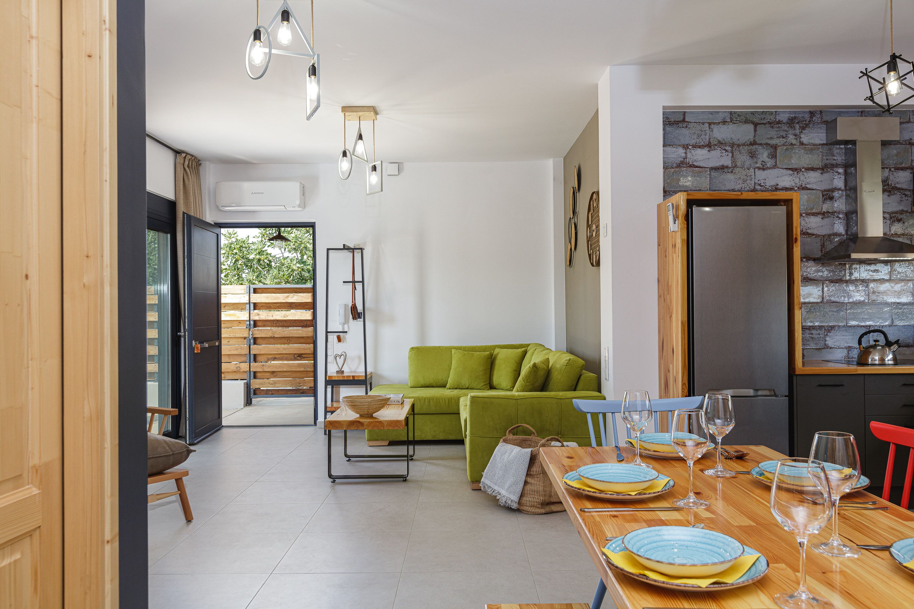 A modern living room with a green sofa, a dining table set with plates and glasses, and a kitchen area with a brick backsplash.