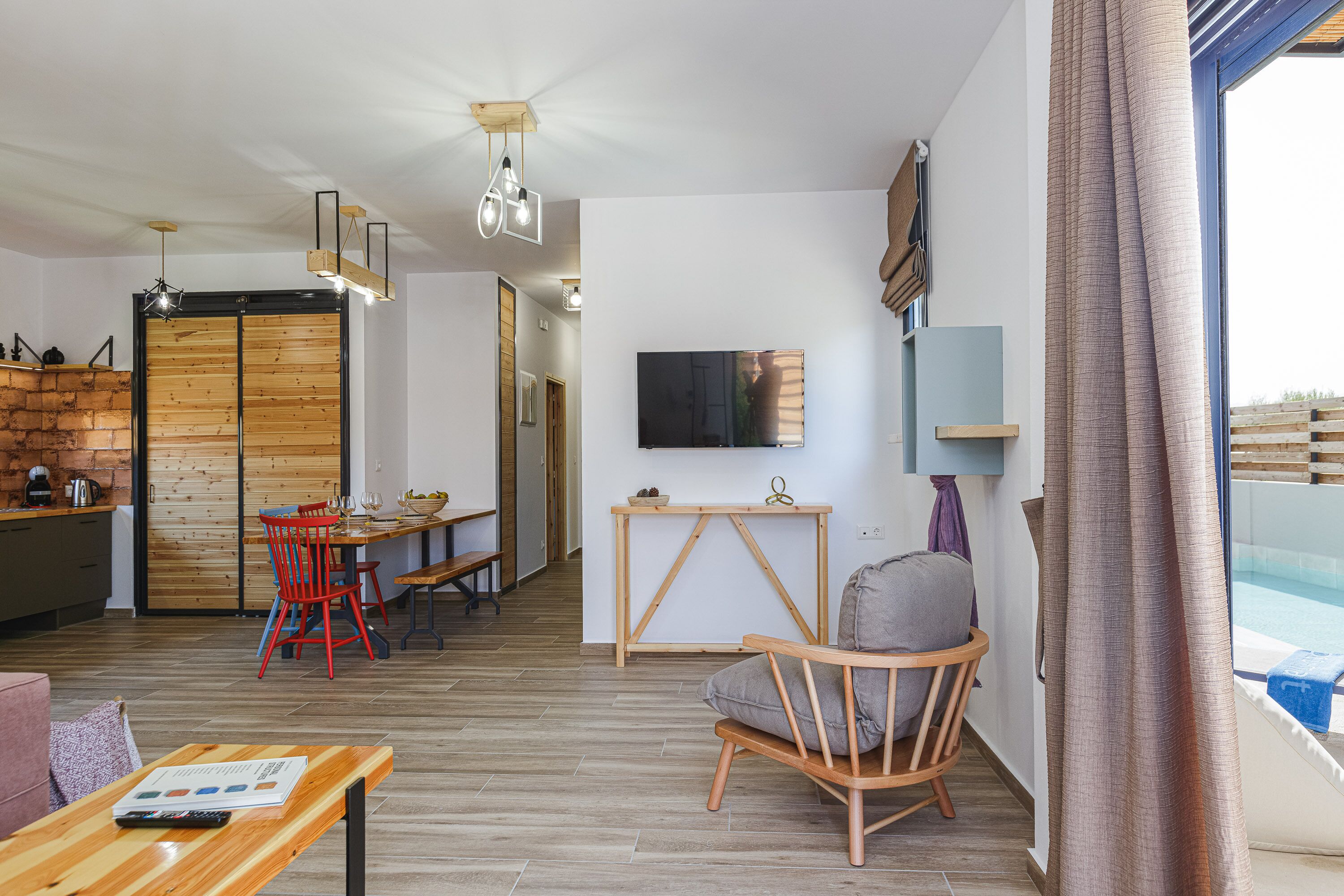 Een moderne woonkamer met een houten vloer, een eethoek met rode stoelen, een aan de muur bevestigde televisie en uitzicht op een zwembad door een raam.