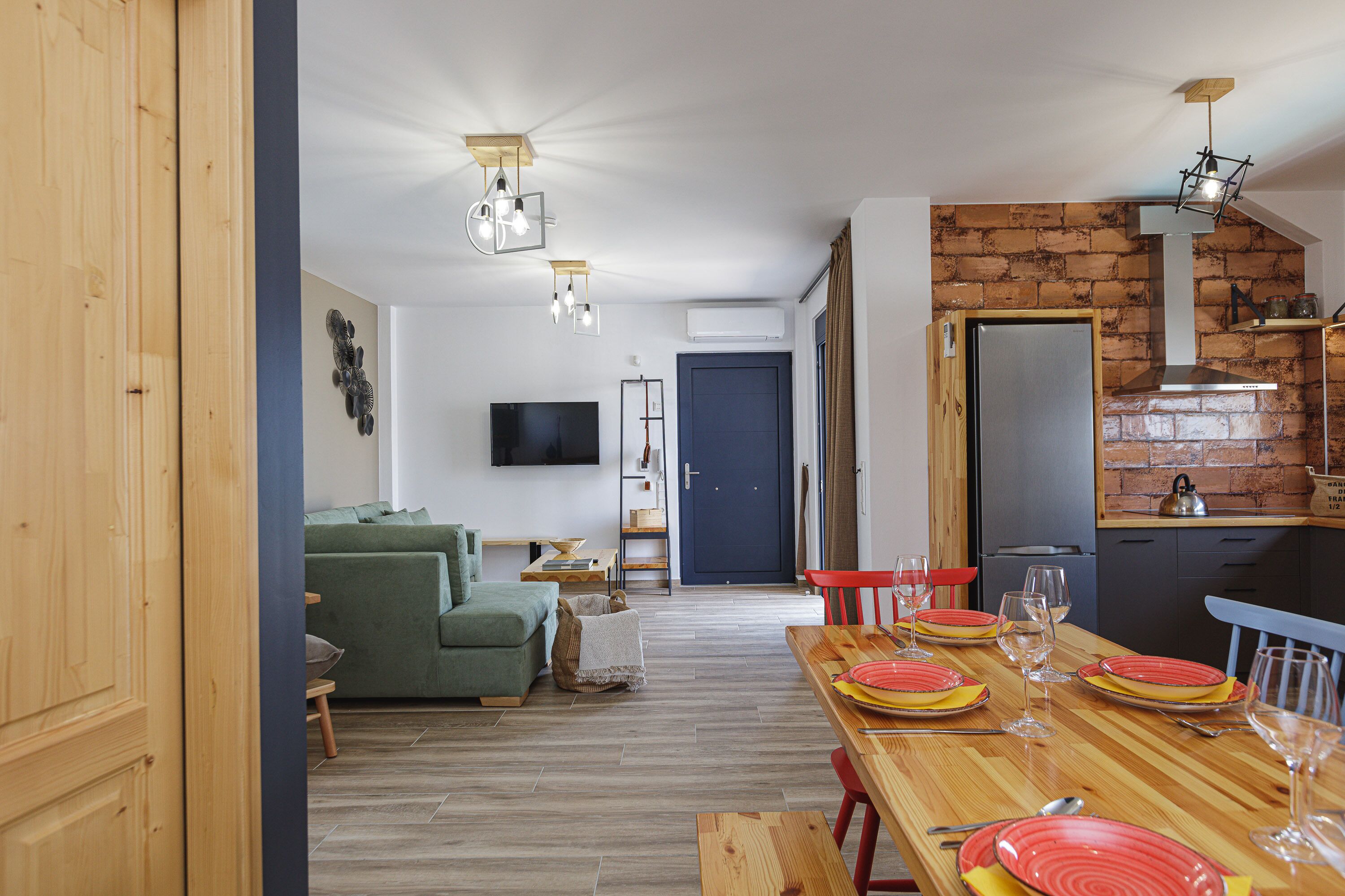 Un soggiorno moderno con un divano verde, un tavolo da pranzo in legno apparecchiato per quattro persone e un angolo cottura con parete in mattoni ed elettrodomestici in acciaio inossidabile.