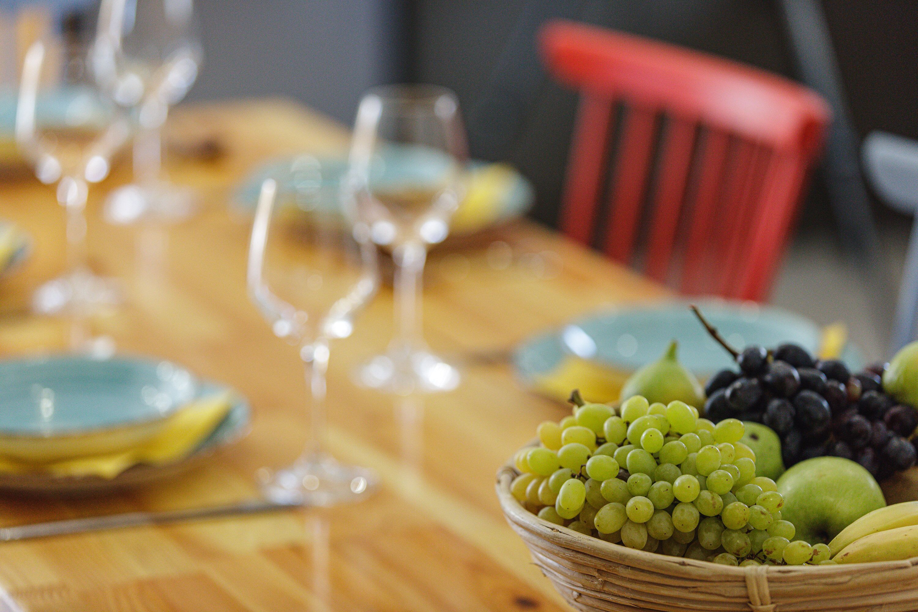A dining table with a wicker basket filled with green grapes, black grapes, and a green apple.
