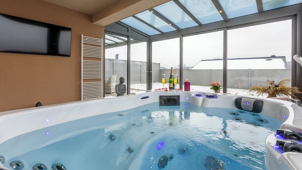 Une piscine intérieure moderne dotée d’un toit en verre, d’un téléviseur à écran plat fixé au mur et d’un jacuzzi avec jets.