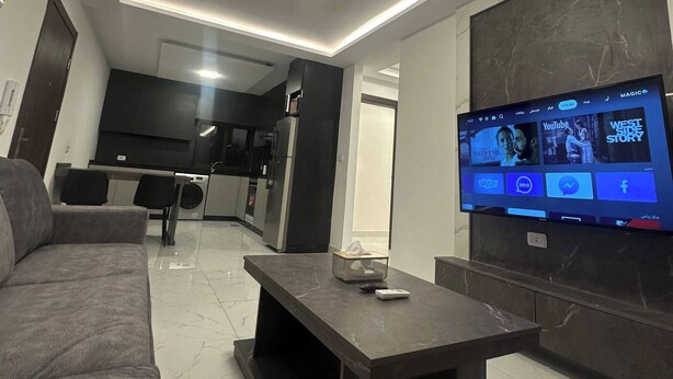 A modern living room with a flat-screen TV, a sofa, a coffee table, and a kitchen in the background. - JRE Cozy Rooftop Near Al Services-23 (Amman)